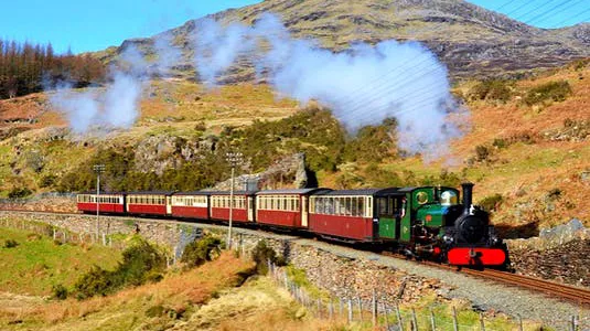 Welsh Highland Railway Steam Train running through the countryside with 4 carriages, with blue skies and steam against a mountain backdrop